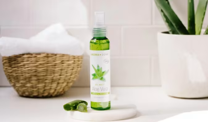 Flacon spray transparent Aroma-Zone Gel Brut Aloe Vera Bio, posé sur une coupelle marbre avec feuille d'aloe vera fraîche, ambiance salle de bain lumineuse avec plante et panier tressé en fond