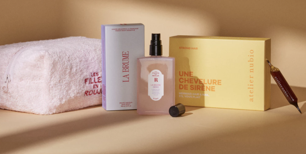 Flacon en verre rose (brume de la marque Rouje), boite en carton jaune (compléments alimentaires "une chevelure de sirène" de la marque Atelier Nubio) à droite et trousse à cosmétiques rose avec inscription "les filles en rouje) à gauche