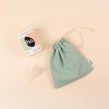 Un kit pour l'hygiène menstruelle éco-responsable posé sur un fond neutre beige rosé. Il comprend une petite boîte en carton portant le logo Jho, une coupe menstruelle en silicone médical transparente pliée, et un pochon de rangement en tissu vert sauge avec un lien coulissant.