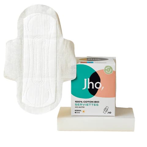 L'image présente une serviette hygiénique blanche déployée, montrant ses ailettes latérales et ses rainures d'absorption. À côté se trouve sa boîte d'emballage blanche et colorée (vert, noir, pêche) marquée "Jho, 100% COTON BIO SERVIETTES". Le tout repose sur un socle blanc épuré