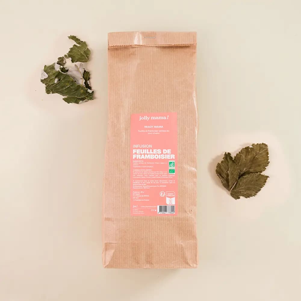 Un sachet en papier kraft brun vertical posé au centre d'un fond crème. L'étiquette rose affiche "jolly mama ! READY MAMA INFUSION FEUILLES DE FRAMBOISIER" avec le logo AB (Agriculture Biologique). Quelques feuilles de framboisier séchées sont disposées artistiquement de chaque côté du sachet.
